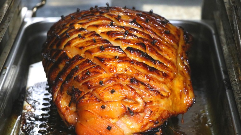 Whole glazed ham roasting on black pan with cross-hatched surface studded with cloves