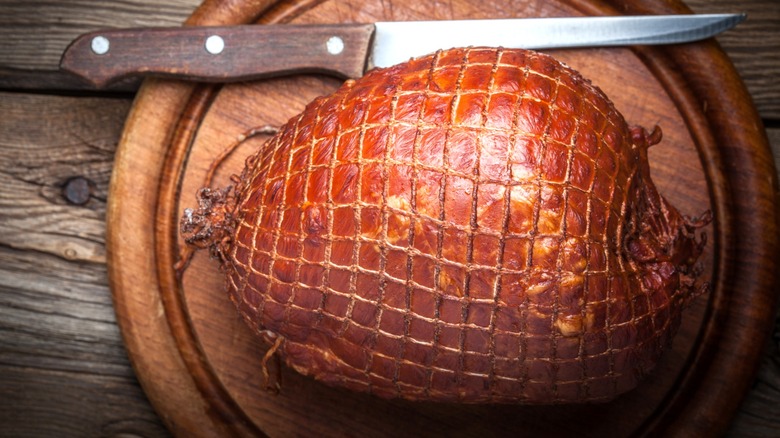 Whole smoked ham in netted casing on round wooden cutting board next to long knife
