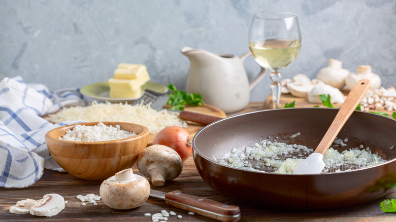 Woman pouring white wine from glass into pan of rice being stirred by chef