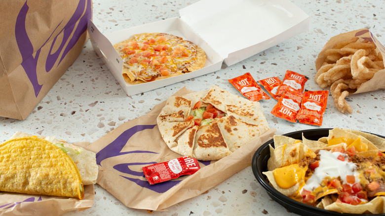 different Taco Bell menu items including a Mexican pizza, a crunchwrap supreme, a hard shell taco, nachos, and cinnamon twists on a table