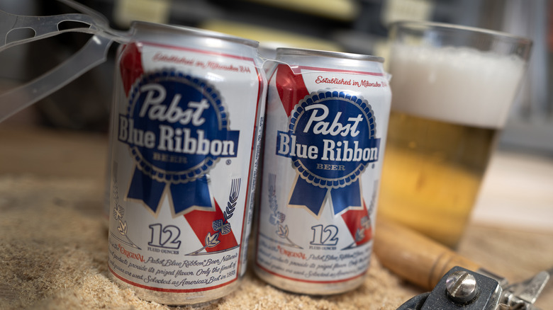 A couple of paps Blue Ribbon beers as part of a six pack, with a glass of beer in the background