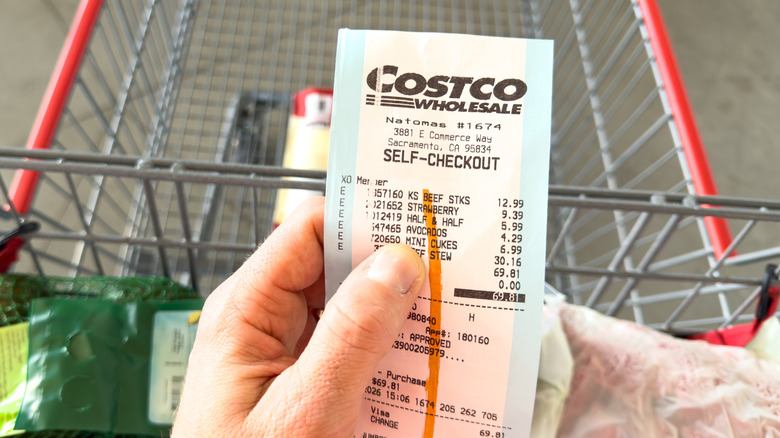 Hand holding checkout receipt from Costco store over shopping cart carrying groceries