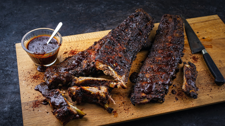 A couple of racks of barbecue ribs on a cutting board scattered with seasoning and sauce
