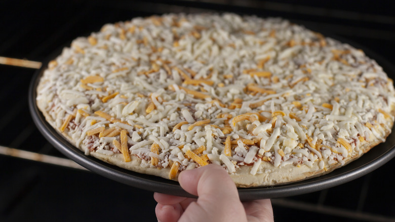 A person is placing a frozen cheese pizza onto an oven rack.
