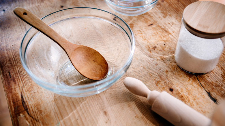 Glass mixing bowl on a wooden countertop with a wooden spoon inside of it next to a jar of white powder and a wooden rolling pin