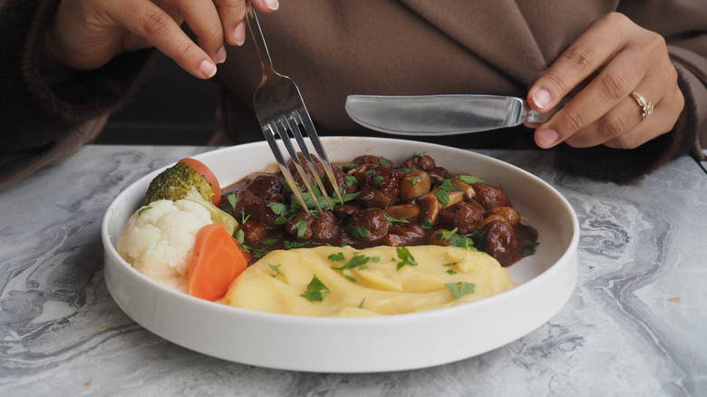 Woman holding a knife and fork to eat beef stew with mashed potatoes.