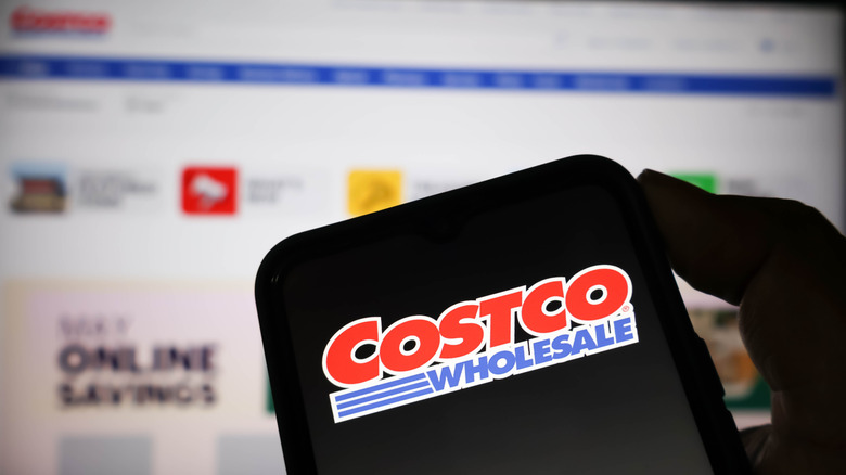 Someone holding a smartphone in front of an out of focus computer screen. The phone displays the name Costco Wholesale