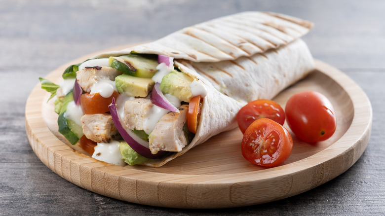 A grilled chicken Caesar wrap cut in half and garnished with cherry tomatoes on a wooden serving board