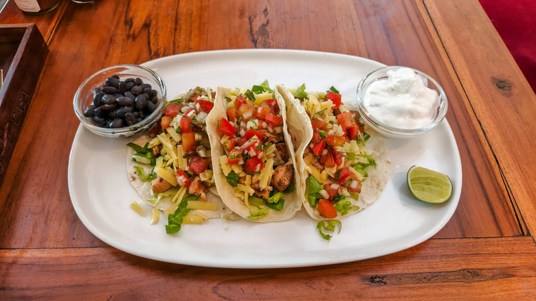 A white platter with three fresh shredded chicken tacos and sides of black beans and sour cream
