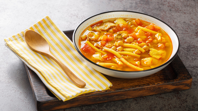 A bowl of hearty tomato soup with pasta and chunks of chicken on a table next to a spoon and napkin