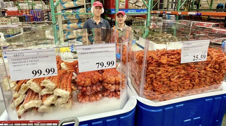 two Costco employees smile at the camera behind displays of king crabs and tiger prawns