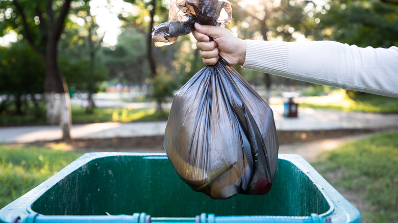 Hand holding a full black trash bag above a green bin