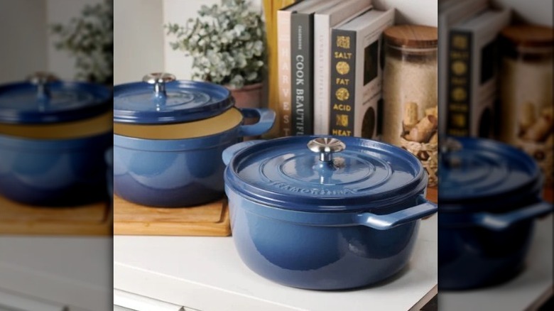 A set of two Tramontina blue enameled cast iron dutch ovens on a kitchen counter with a plant and cookbooks behind them.