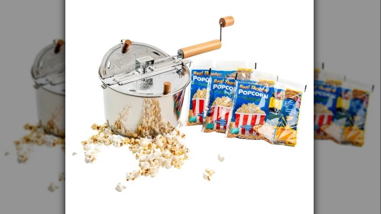 A beauty shot of the Whirley Pop stainless steel popcorn popper, three bags of popcorn kernels, and fresh popped corn scattered on a white backdrop.