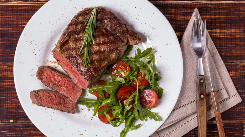 A grilled ribeye steak with two slices removed, topped with fresh rosemary, and sitting on plate alongside an arugula and tomato salad.
