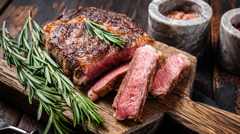 A cooked and sliced ribeye steak resting on a wooden chopping board alongside some fresh rosemary.