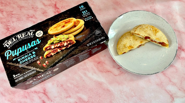Del Real birria cheese pupusa cut in half on white plate next to box, on pink marble surface