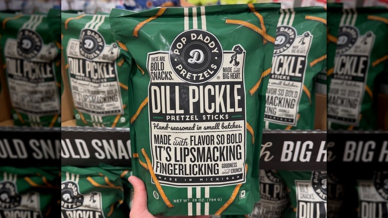 A hand holding a green bag of Pop Daddy Pretzels Dill Pickle Pretzel Sticks in front of a display at Costco