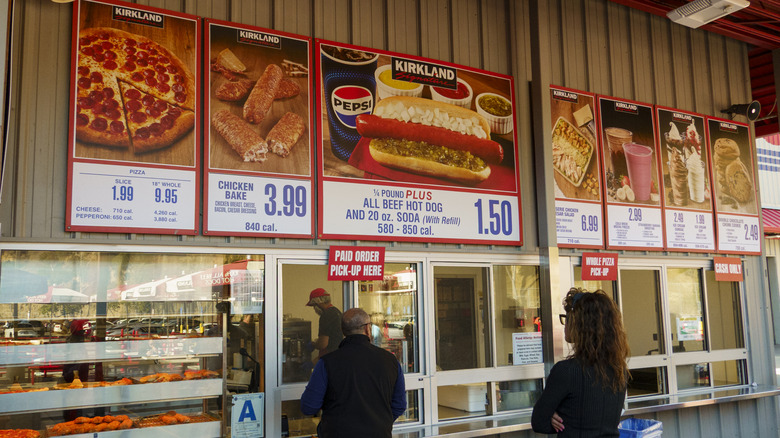A Costco food court with people lined up at the window and menu boards across the wall