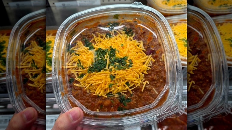 A hand holding a container of Costco beef chili topped with shredded cheese