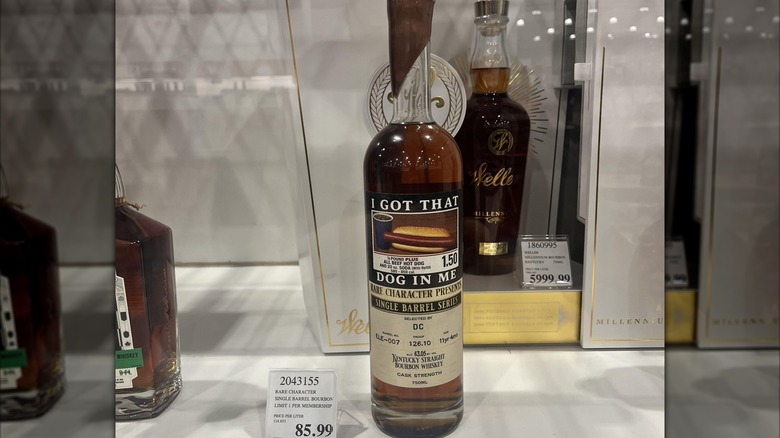A bottle of bourbon labeled "I Got That Dog in Me," with an image of a Costco hot dog on it, sitting on a shelf next to a price tag that says $85.99