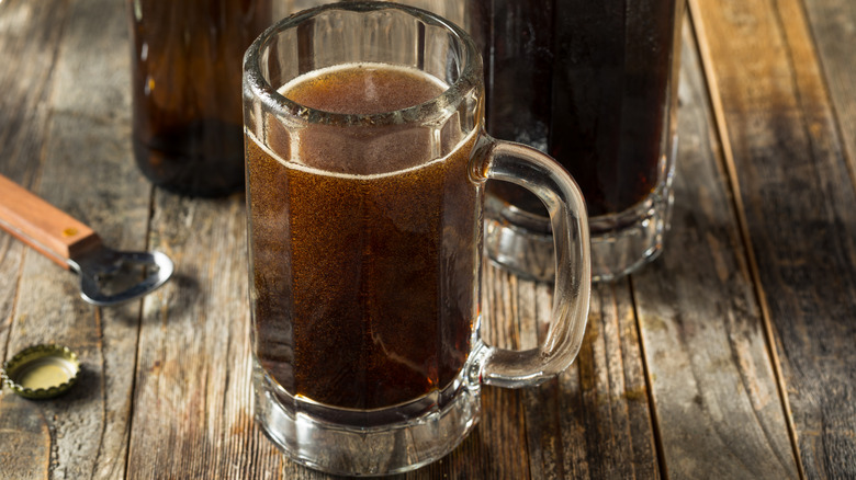 Two glass mugs of fizzy brown soda on a wooden table with a brown glass bottle and bottle opener