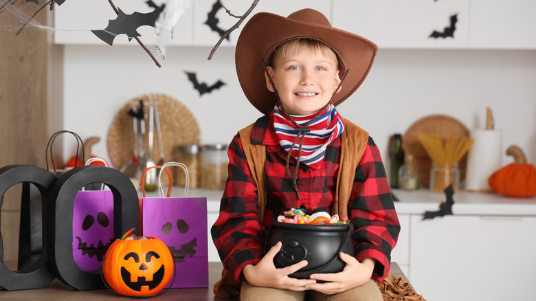 Little boy dressed as cowboy for Halloween with candies on table in kitchen