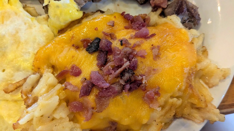 A close-up of a pile of hashbrowns covered in cheese and fresh bacon bits on a white plate next to eggs