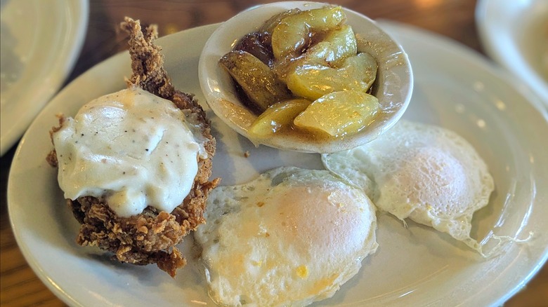 Two over-easy eggs, a piece of fried chicken, and a bowl of fried apples on a white plate