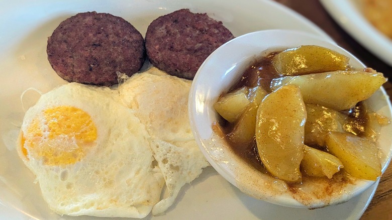 Two fried eggs, two small sausage patties, and a bowl of fried apples on a white plate