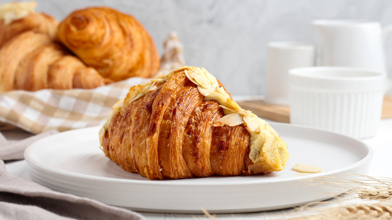 Almond croissants on a white plate with others behind on checkered kitchen towel