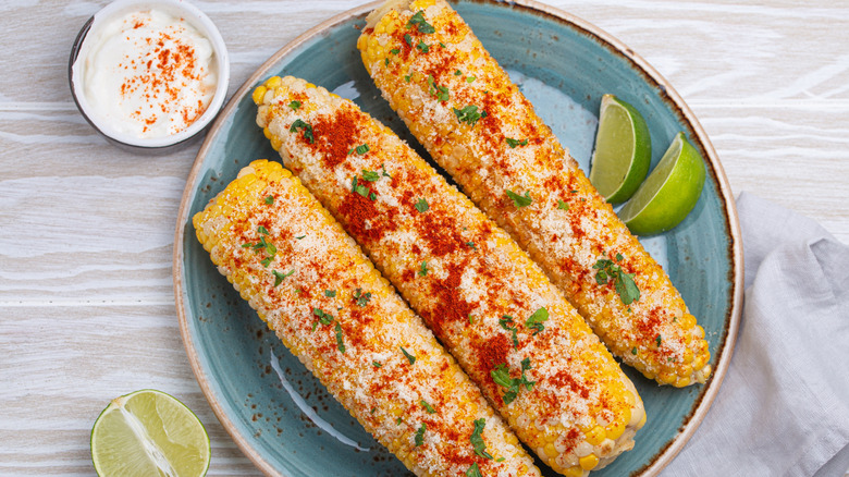 home prepared elote on a plate with lime wedges and mayo or crema besides