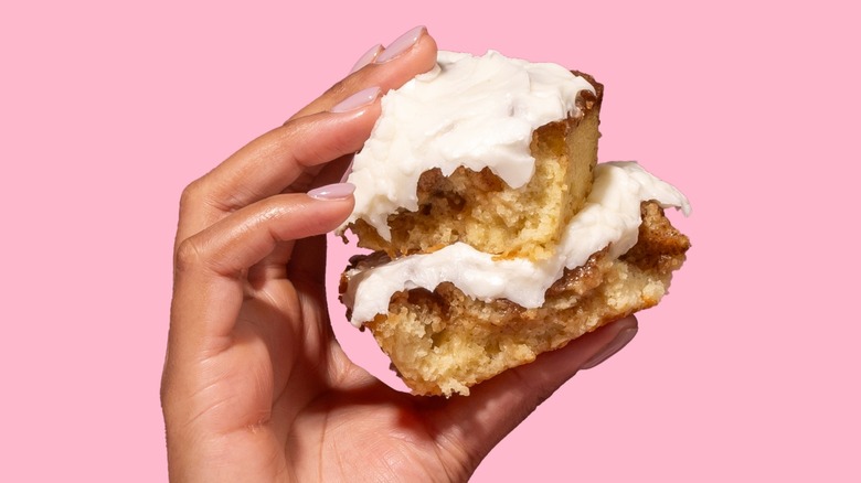 A hand holding a Crumbl Cinnamon Square with a pink background