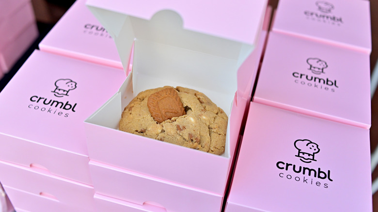 Stacks of small, square pink Crumbl boxes with the top one open, showcasing a cookie.