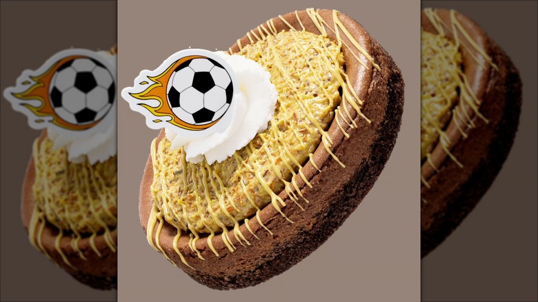 A product image of Crumbl's Dubai-style cheesecake cookie, topped with whipped cream and a soccer ball topper.