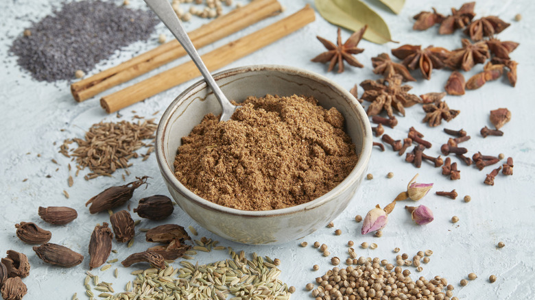 Small bowl of garam masala with a spoon in it surrounded by the whole spices