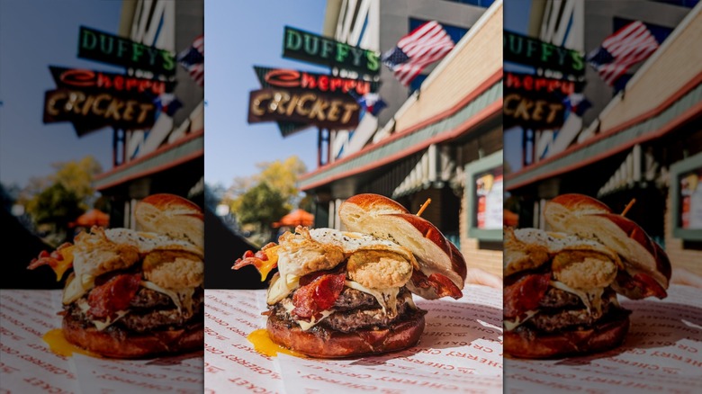 A loaded burger from The Cherry Cricket on a table in front of the resturant's outdoor sign