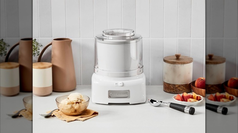 White Cuisinart Ice Cream Maker on white counter next to bowl of ice cream, ice cream scoop, and various jars and pitchers