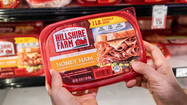 A hand holds a package of Hillshire Farm Honey Ham in a grocery aisle.