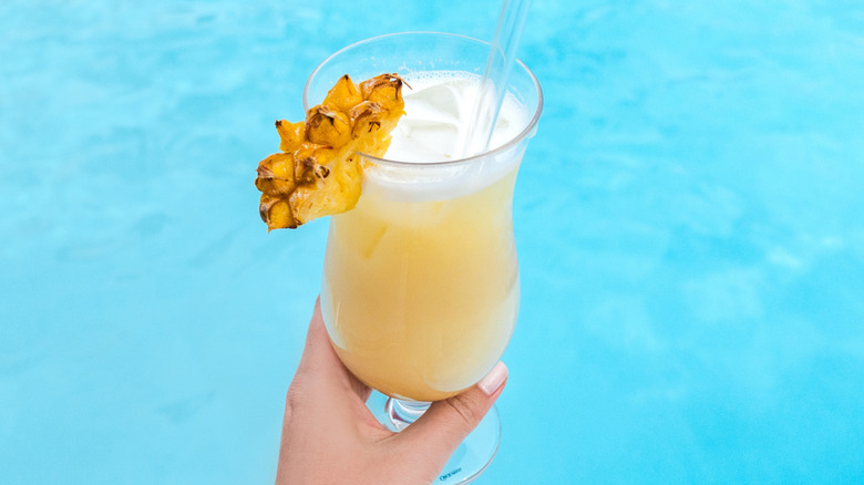Person holding tropical cocktail poolside.