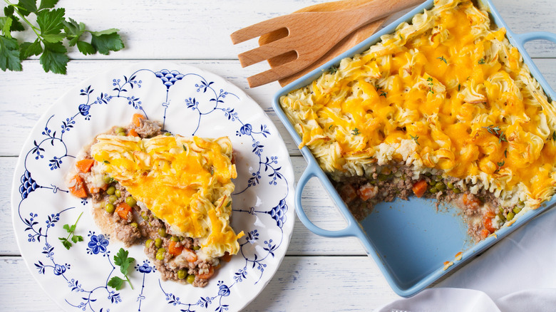 shepherd's pie with cheese topping shown in a square baking dish and a portion on a plate all on a wooden table top.