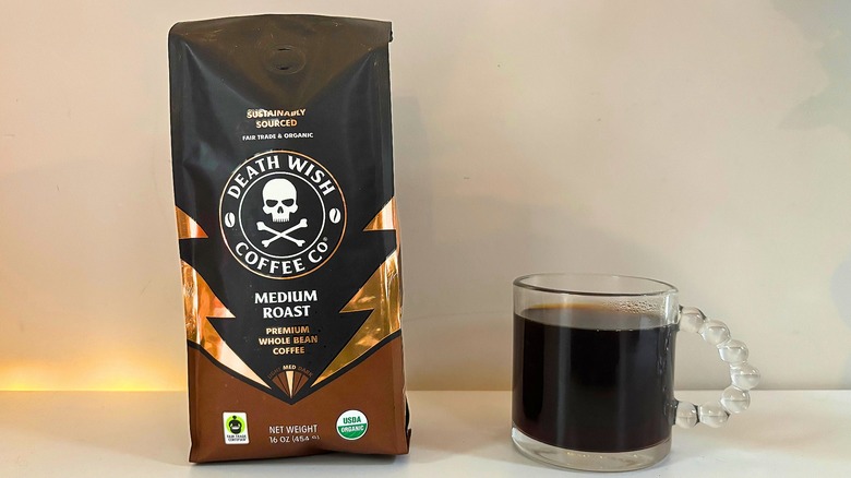 Bag of Death Wish Medium Roast coffee next to a clear mug of black coffee on a white table