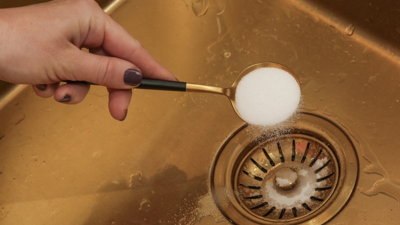 Hand pouring a spoonful of white powder into a kitchen sink drain