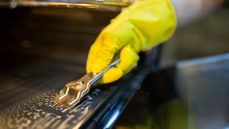 A person wearing yellow rubber gloves scraping grime off of an oven using a metal razor scraper tool