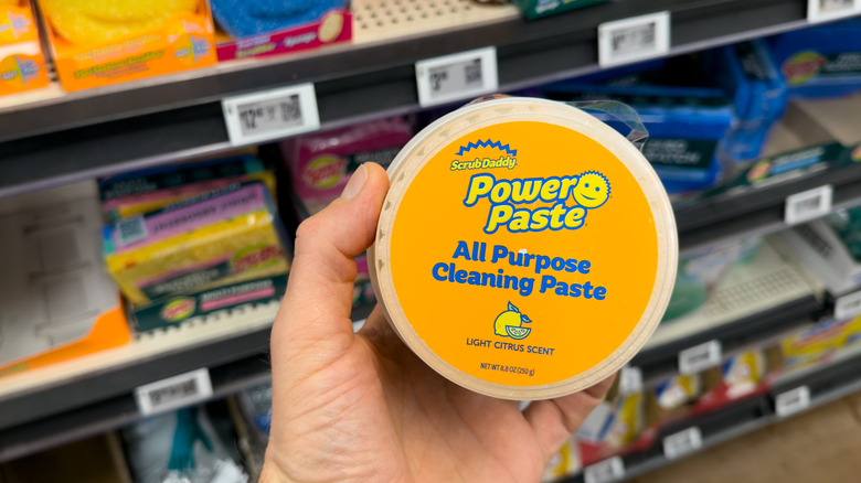 A hand holding a yellow and blue container of Scrub Daddy Power Paste in a grocery store