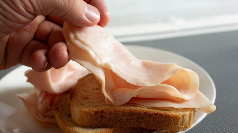A person places a slice of pink deli meat on a stack of dry bread