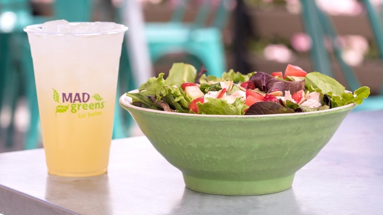 Salad in a green bowl and a pale yellow drink in a MAD Greens cup beside it