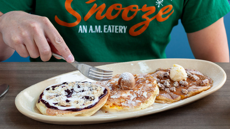 Person wearing a Snooze T-shirt holding a fork above plate of pancakes