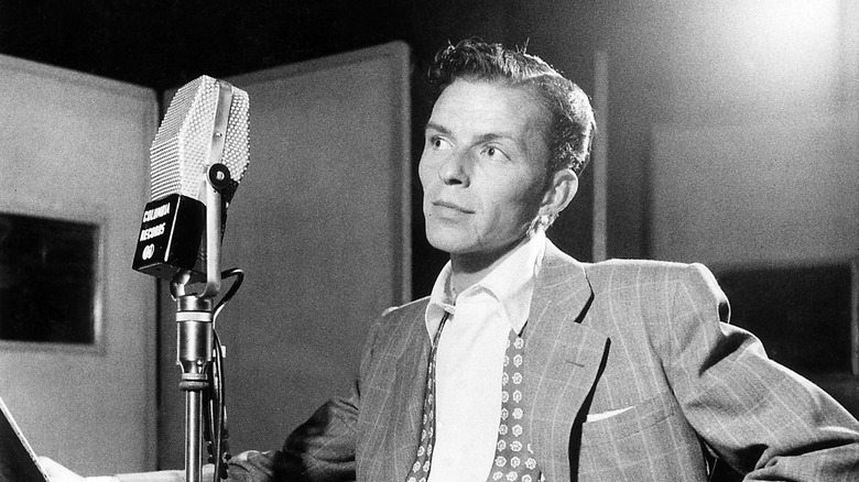 A black-and-white photograph of a young Frank Sinatra standing in front of a microphone in a recording studio