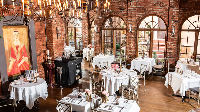 A restaurant dining room with wooden floors and red brick walls featuring tables adorned with white tablecloths and flower centerpieces with chandeliers hanging from the ceiling and a painting of a woman in a red dress on the wall